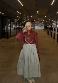 Femme portant un foulard beige, un haut bordeaux et une jupe vert clair, debout sous un plafond éclairé dans un espace intérieur spacieux.