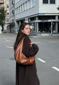 Pruun nahast õlakott, mis on pehme ja mahukas ning millel on suur avaus. Kandmise all must ülegrootud mantel, linnatänava taustal.