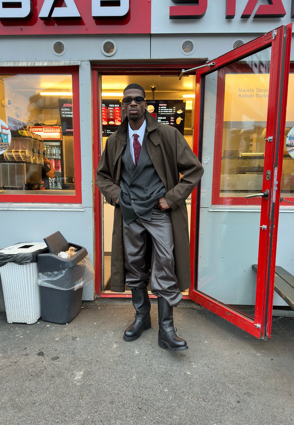 Uomo con occhiali da sole, cappotto trench, completo e stivali in piedi nella porta di un negozio di kebab con porta rossa aperta.