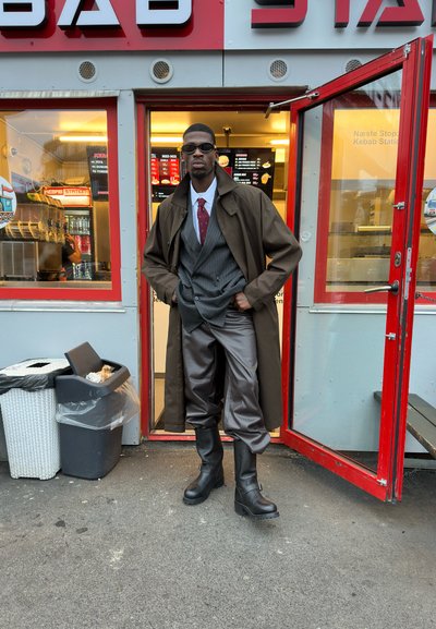 Hombre con gafas de sol, gabardina, traje y botas de pie en la puerta de un kebab con la puerta roja abierta.