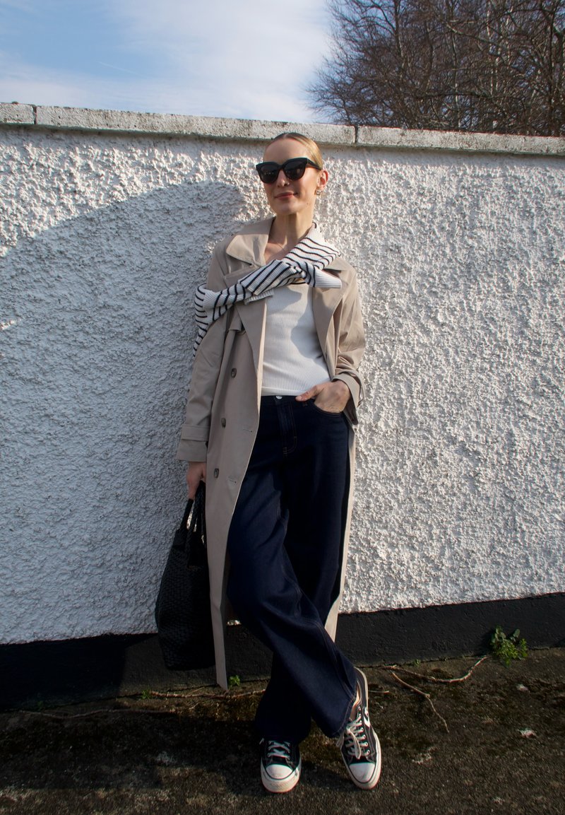 Mulher vestindo casaco trench coat bege, top branco, calças largas escuras, suéter às riscas sobre os ombros, óculos de sol pretos, segurando mala preta, em pé junto a uma parede.