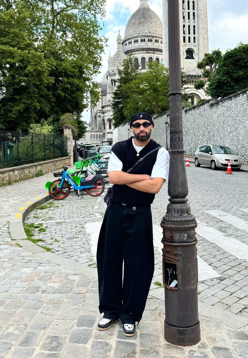 Μαύρο παντελόνι φαρδύ και μαύρο αμάνικο πουλόβερ πάνω από λευκό πουκάμισο. Αξεσουάρ περιλαμβάνουν ένα μαύρο μπερέ και γυαλιά ηλίου. Φόντο με πλακόστρωτο δρόμο.