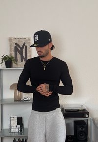 Un uomo che indossa una maglietta nera a maniche lunghe, pantaloni da tuta grigi e un cappellino nero si trova davanti a degli scaffali con piante, opere d'arte, libri e attrezzature audio.