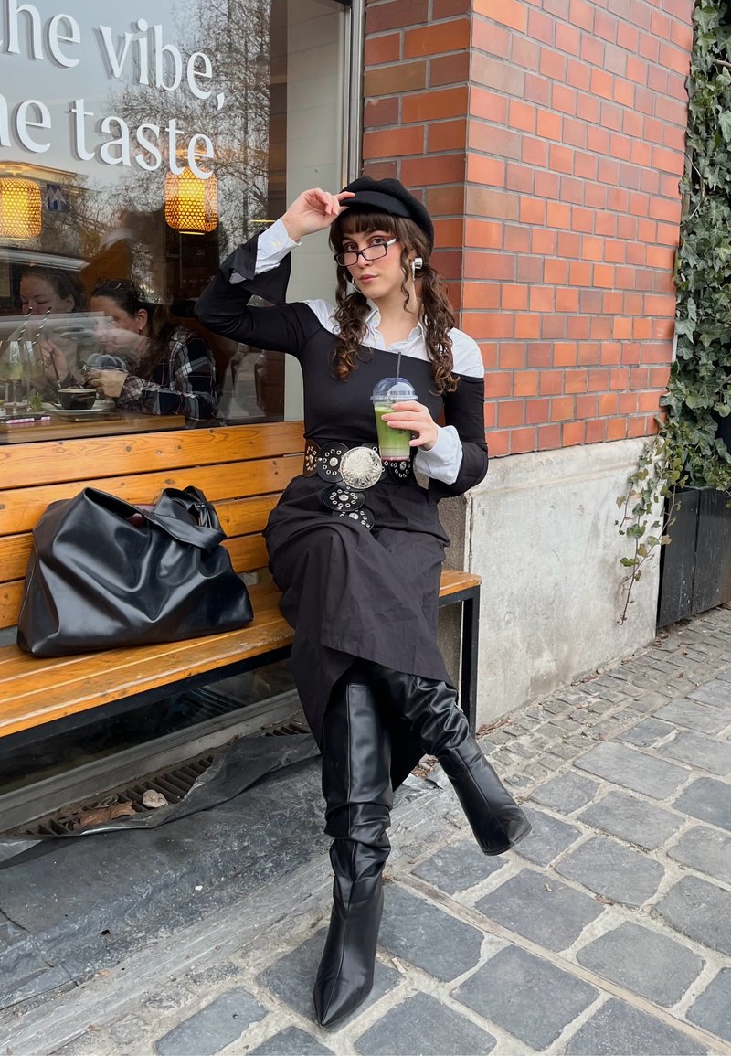Femme portant un béret noir, des lunettes et des bottes, assise sur un banc à l'extérieur d'un café, tenant une boisson verte, un sac noir à ses côtés, mur en briques en arrière-plan.