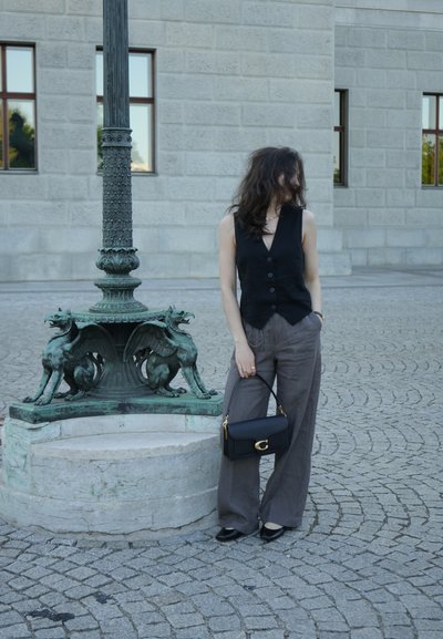 Chaleco negro sin mangas con botones, pantalones de pierna ancha gris y un bolso negro con un acento de hardware dorado, de pie cerca de un pilar de piedra con grifos.