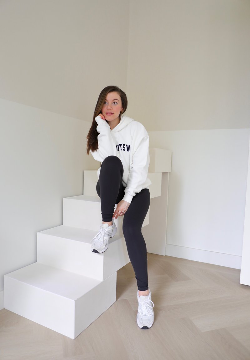 Λευκή φόρμα με κουκούλα και ναυτικές λεπτομέρειες, μαύρες κολάν και γκρι αθλητικά παπούτσια. Το άτομο κάθεται σε λευκά γεωμετρικά σκαλοπάτια με ξύλινο δάπεδο.