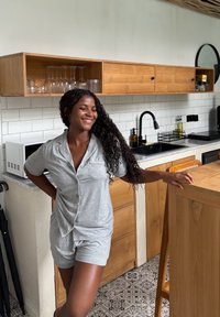 Femme en pyjama gris souriante s'appuyant sur un îlot de cuisine en bois dans une cuisine moderne avec des verreries et un évier en arrière-plan.