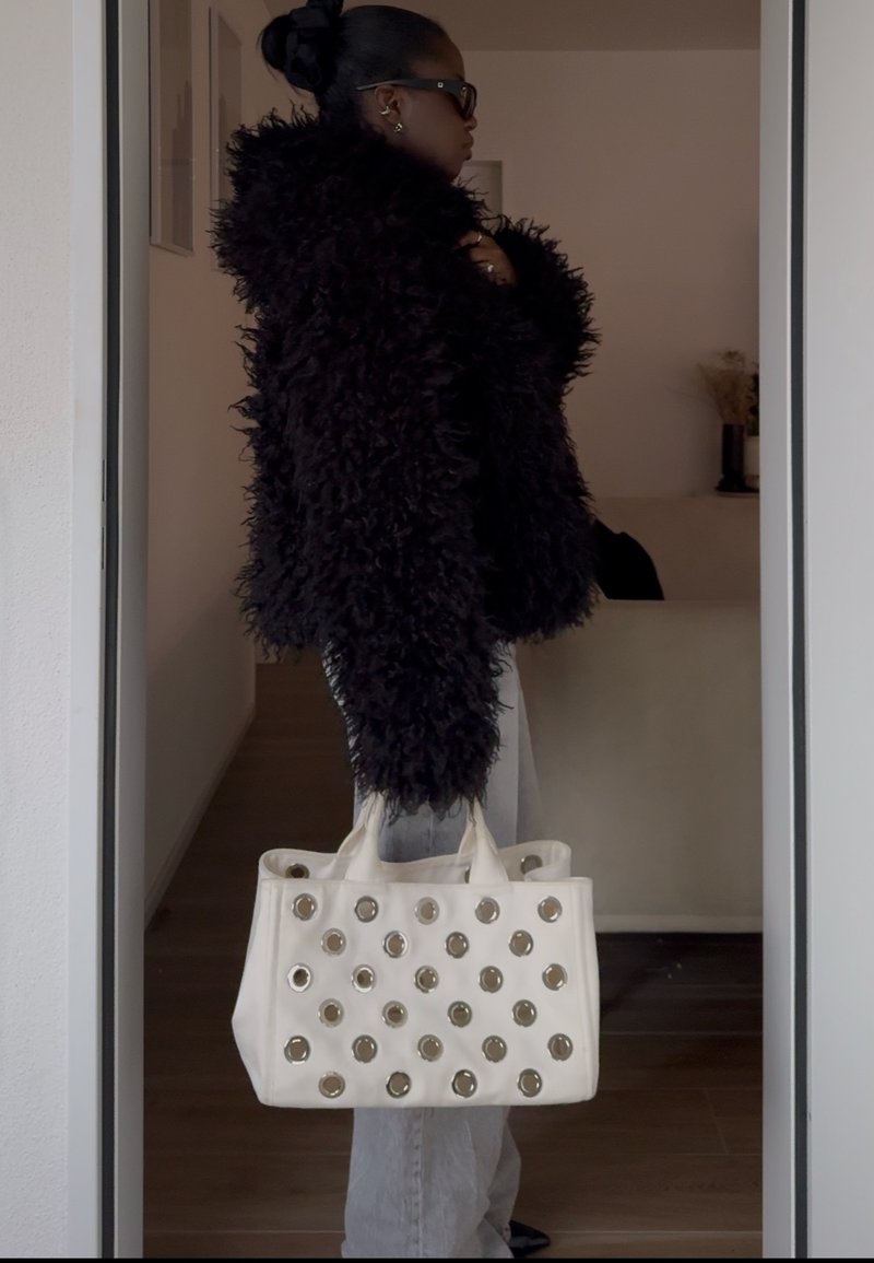 Femme en manteau noir duveteux et lunettes de soleil tenant un sac à main blanc avec des détails métalliques circulaires, se tenant à l'intérieur près d'une porte.