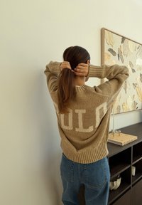 Γυναίκα με καστανά μαλλιά, ντυμένη με μπεζ πουλόβερ και μπλε τζιν, δένει τα μαλλιά της σε μια χαμηλή αλογοουρ ά κοντά σε σκούρα ράφια και αφηρημένη τοιχογραφία.