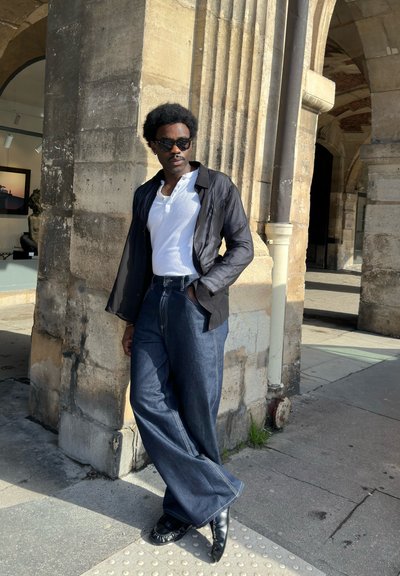 Hombre con gafas de sol, camisa blanca, chaqueta negra y pantalones anchos azules apoyado contra un arco de piedra en un entorno urbano soleado.