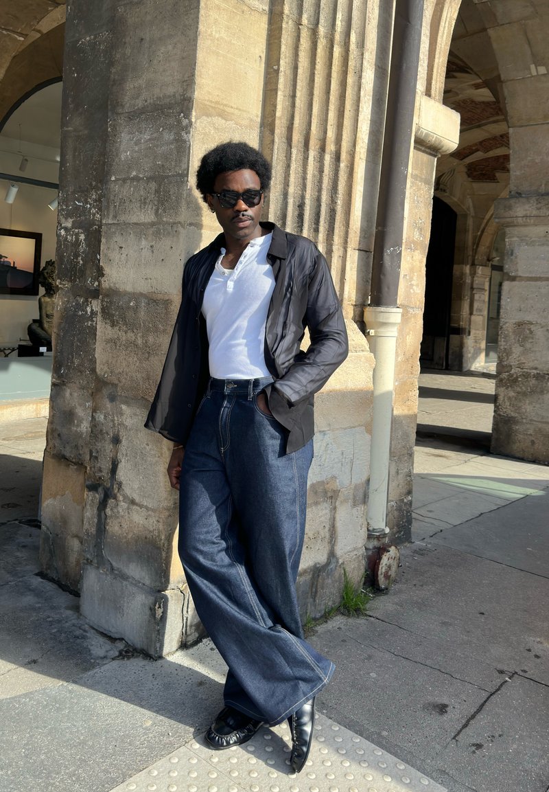 Homme portant des lunettes de soleil, chemise blanche, veste noire et jean bleu large, appuyé contre une arche en pierre dans un cadre urbain baigné de lumière.