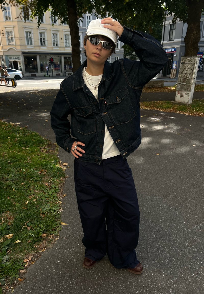 Person som har på seg mørk denimjakke, vide svarte bukser, hvit caps, solbriller og ringer, står på en urban fortau med trær og bygninger i bakgrunnen.