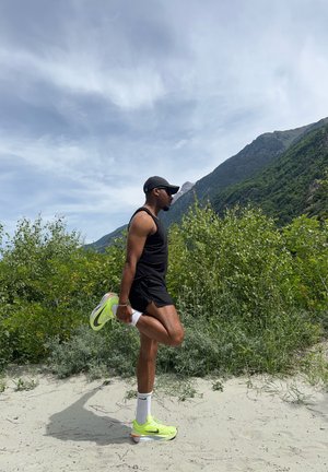 Scarpe sportive di un vivace giallo neon con suole bianche, pantaloni corti neri e una canotta nera senza maniche. Lo sfondo presenta vegetazione e montagne.