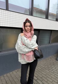 Vastag szürke kötött pulóver, rózsaszín bojtokkal díszített sál, fekete kézitáska, fekete nadrág, modern, csempézett ablakú épület előtt állva.