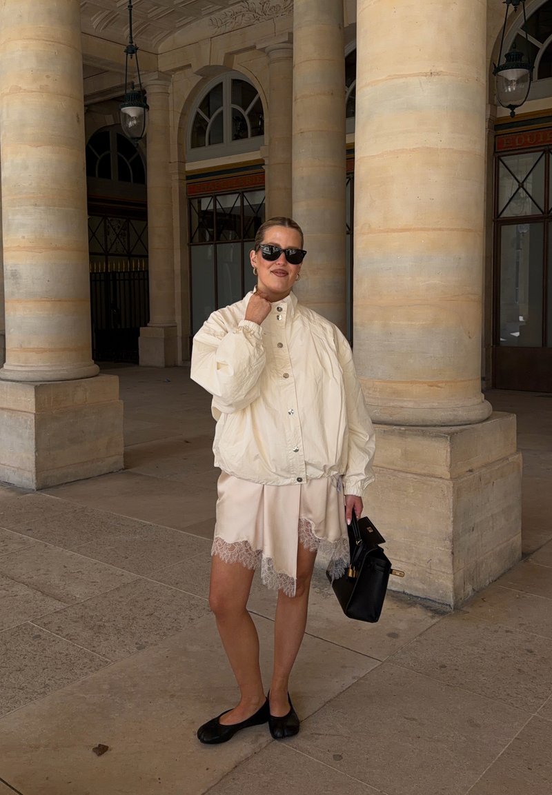 Femme en veste crème, jupe beige bordée de dentelle, ballerines noires et lunettes de soleil tenant un sac à main noir, debout près de colonnes en pierre.