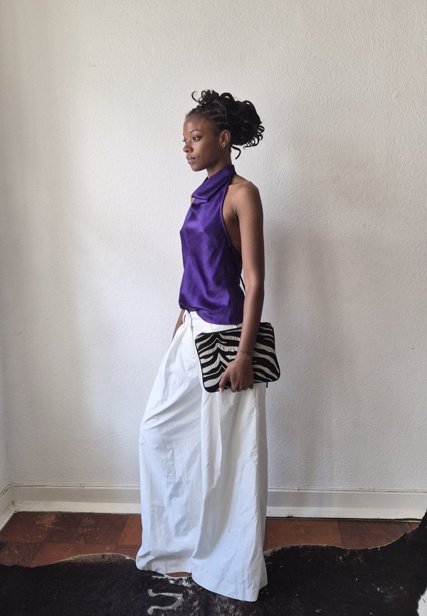 Une femme portant un chemisier violet sans manches et une longue jupe blanche tient une pochette à motifs noirs et blancs et se tient devant un mur uni.