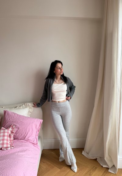 Mujer con cárdigan gris, top corto blanco y pantalones grises apoyada en una cama con ropa de cama rosa, de pie cerca de una cortina crema y una pared beige.