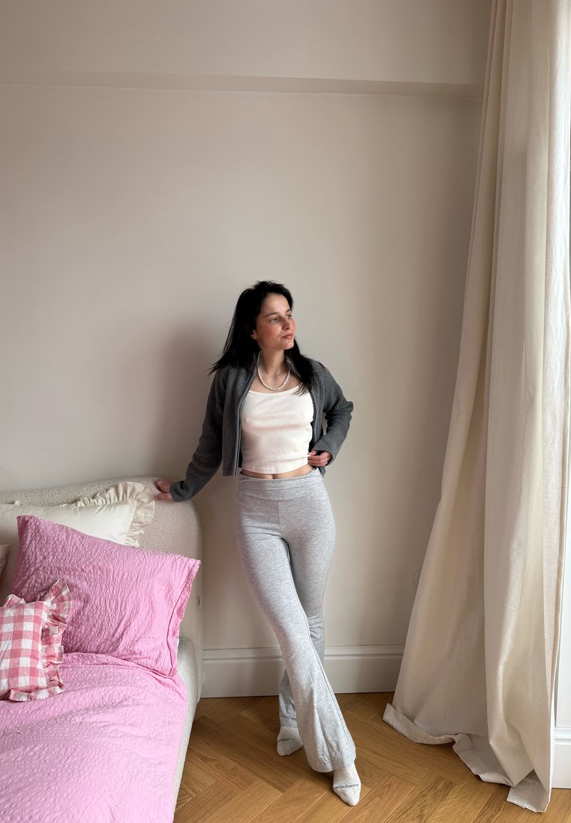 Femme en cardigan gris, haut court blanc et pantalon gris, appuyée sur un lit avec une literie rose, debout près d'un rideau crème et d'un mur beige.