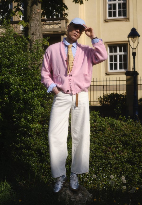 Osoba koja nosi svijetloplavu kapu, ružičasti kardigan, žutu kravatu i bijele široke hlače stoji na stijeni u vrtu ispred zgrade.