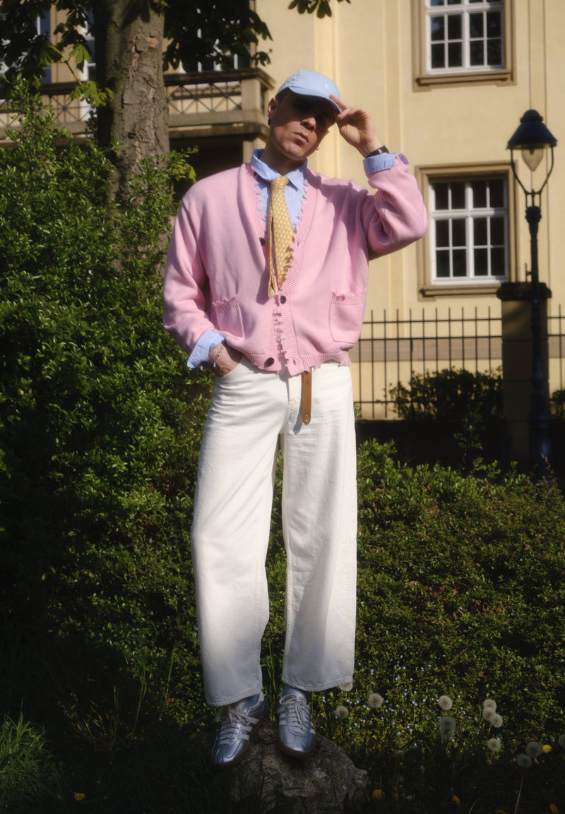 Personne portant une casquette bleu clair, un cardigan rose, une cravate jaune et un pantalon large blanc debout sur un rocher dans un jardin devant un bâtiment.