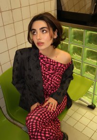 Geruite roze en zwarte jurk met drapering, gecombineerd met een donker blazer met een bloemenpatroon. Zittend op een groene stoel.