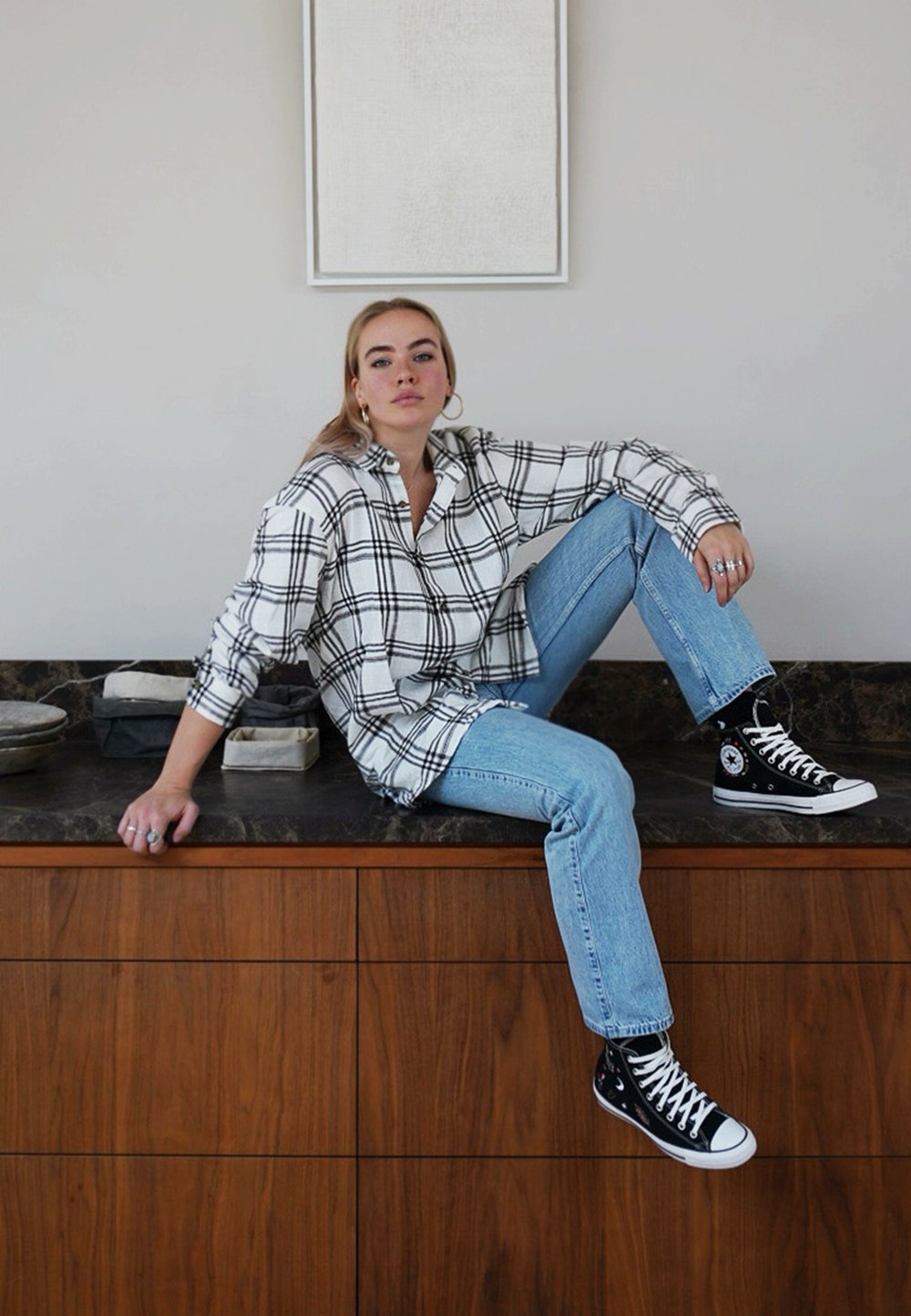 Camisa xadrez em preto e branco, jeans azul claro, sapatilhas pretas de cano alto, sentado sobre uma superfície de madeira com recipientes de armazenamento cinza e pretos.