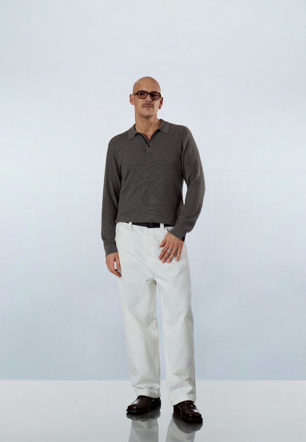 Man draagt een donkergrijs shirt met lange mouwen, witte broek, zwarte riem, donkere schoenen en bril, staat op een reflecterend oppervlak tegen een lichte achtergrond.