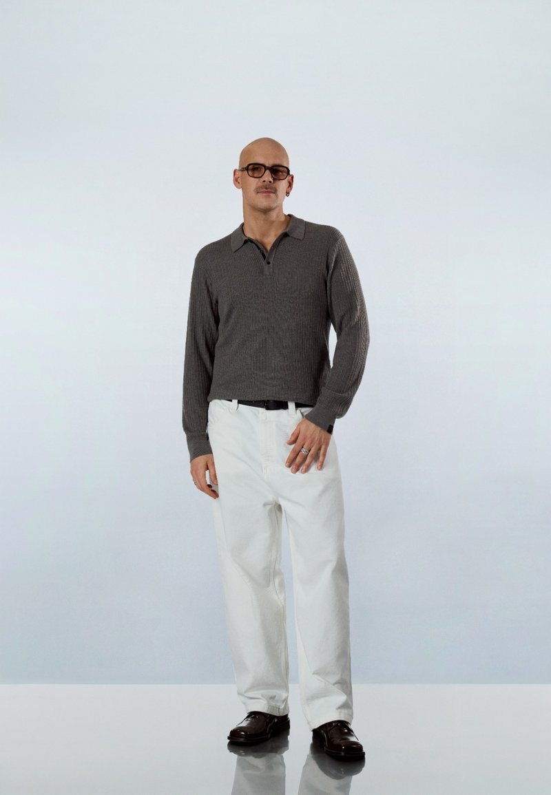 Uomo che indossa una maglia a maniche lunghe grigio scuro, pantaloni bianchi, cintura nera, scarpe scure e occhiali, in piedi su una superficie riflettente con sfondo chiaro.