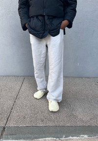 Μαύρο μπουφάν, λευκά λινά παντελόνια και ανοιχτά κίτρινα ξύλινα υποδήματα. Τα παντελόνια είναι φαρδιά και έχουν μια λεία υφή σε γκρι φόντο.