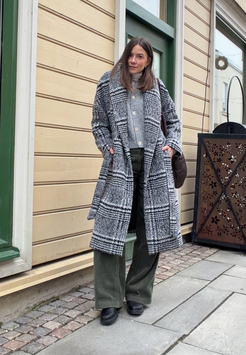 Lang ternet frakke i sort og hvid, tekstureret uldblanding. Grå strikket sweater med knapper, olivengrønne vid-ben fløjlsbukser, sorte støvler.