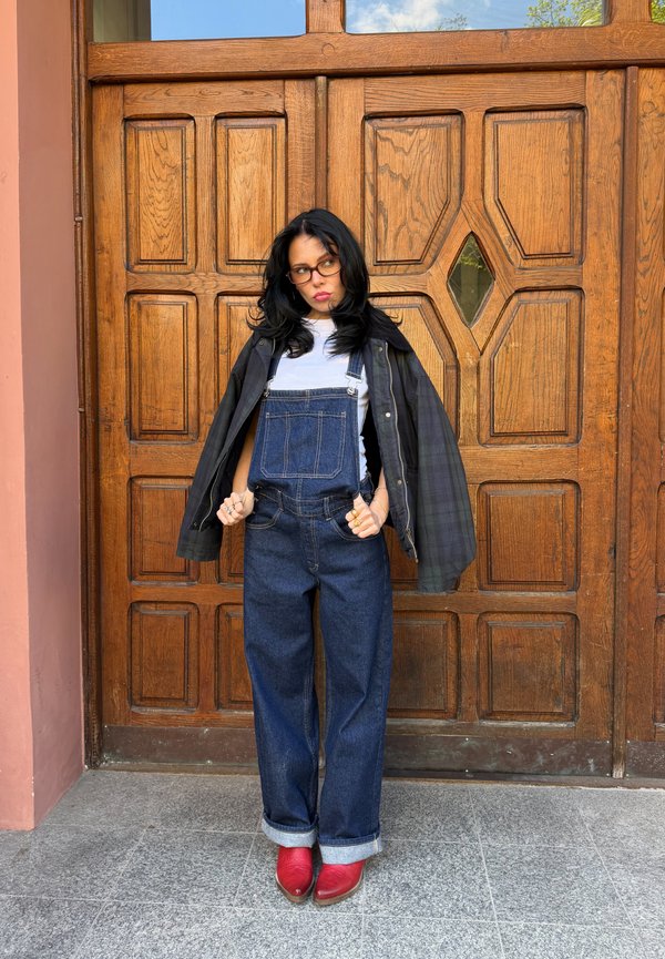 Joven mujer con cabello negro y gafas que lleva un peto vaquero, camisa blanca, chaqueta negra y zapatos rojos, está de pie junto a una puerta de madera.