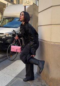 Schwarze Kunstfelljacke, schwarze lockere Hose, schwarze Gummistiefel und eine pinke Handtasche. Der Hintergrund enthält ein Fahrrad und ein Lieferfahrzeug.