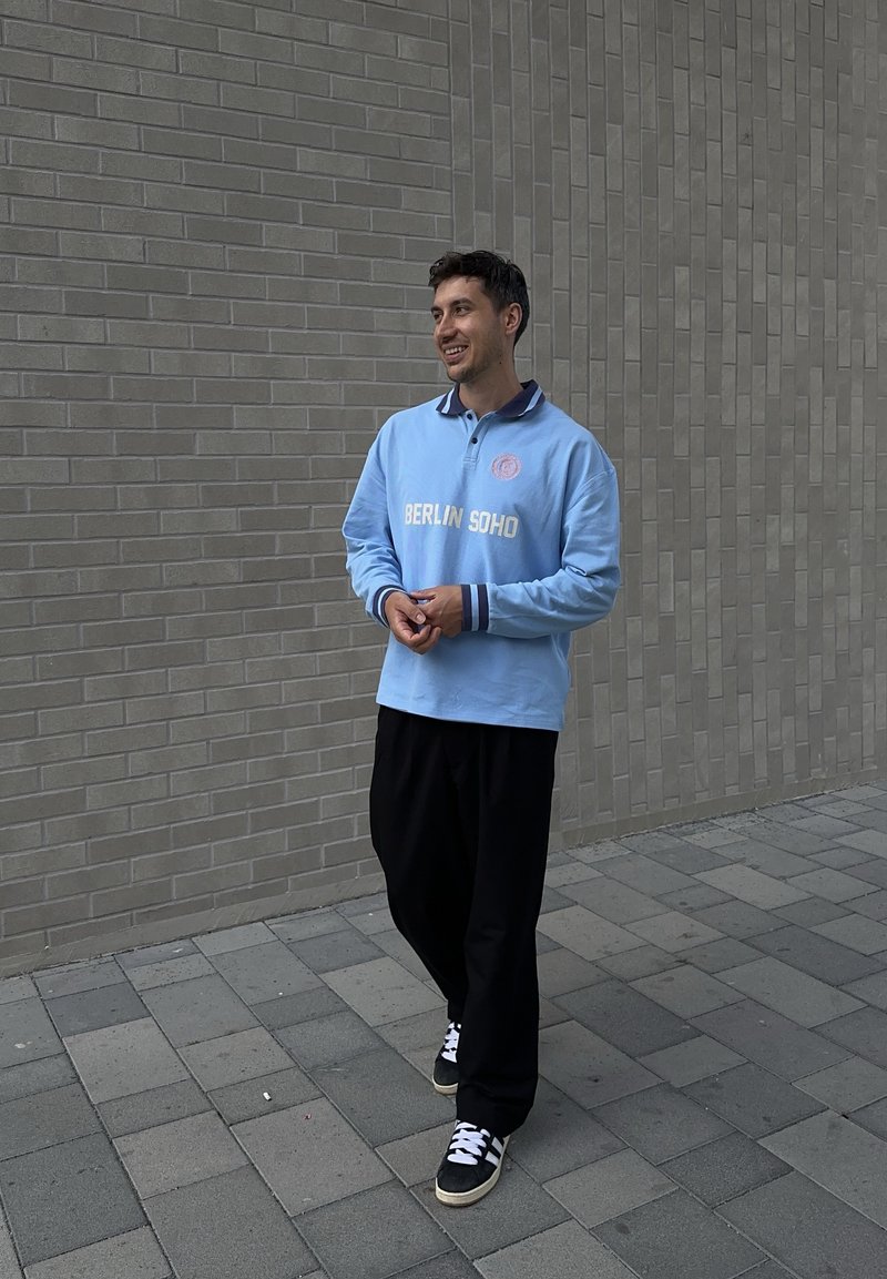 Poloshirt in Hellblau mit navyfarbenem Kragen und Bündchen, versehen mit dem weißen Text "BERLIN SOHO"; kombiniert mit lockeren schwarzen Hosen und dunklen Sneakern.