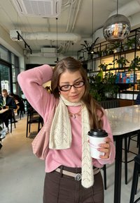 Un pull tricoté rose clair, une écharpe en dentelle crème, un pantalon marron avec une boucle de ceinture en forme de rose, tenant une tasse de café noir dans un café avec des plantes.