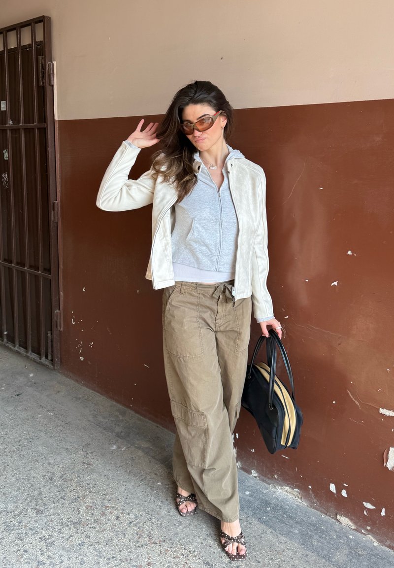 Γυναίκα με γυαλιά ηλίου φορώντας λευκό μπουφάν, γκρι φούτερ με φερμουάρ, φαρδιά καφέ παντελόνια και σανδάλια, κρατώντας μαύρη τσάντα ενώ στέκεται δίπλα σε καφέ τοίχο.