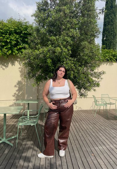 Pantalones de símil cuero marrón de pierna ancha y una blusa corta blanca, de pie en una terraza de madera con muebles metálicos verdes y arbustos de fondo.