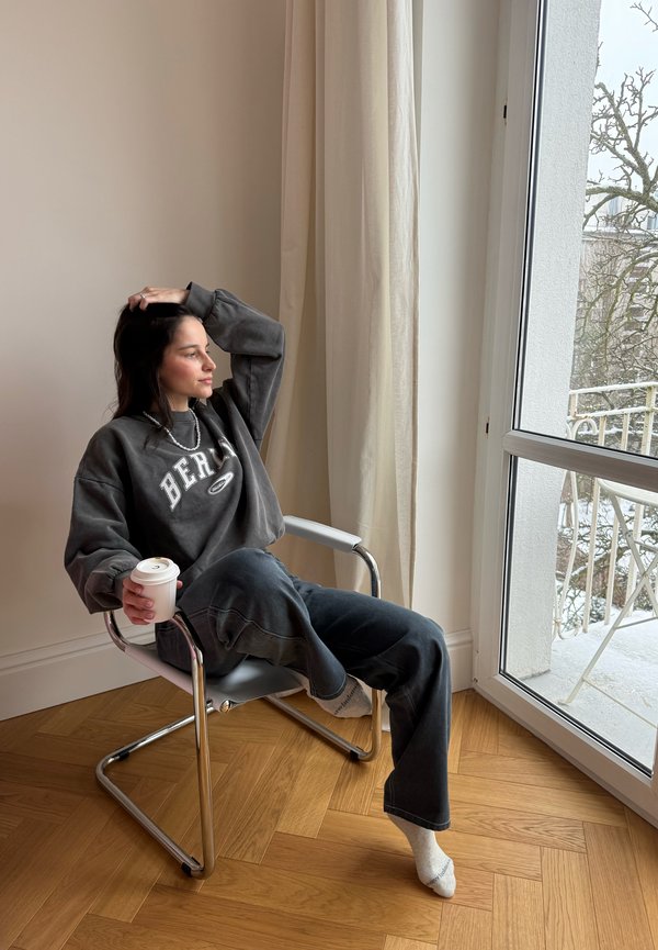 Nainen rentoihin vaatteisiin pukeutuneena istuu tuolilla ikkunan vieressä, pitelee kahvikuppia ja katsoo ulos, toinen käsi lepää hänen päänsä päällä.