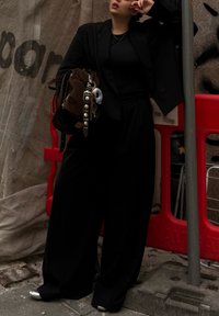 Black oversized blazer worn over a black top, teamed with wide black trousers. Accessories consist of a brown bag featuring metallic accents.