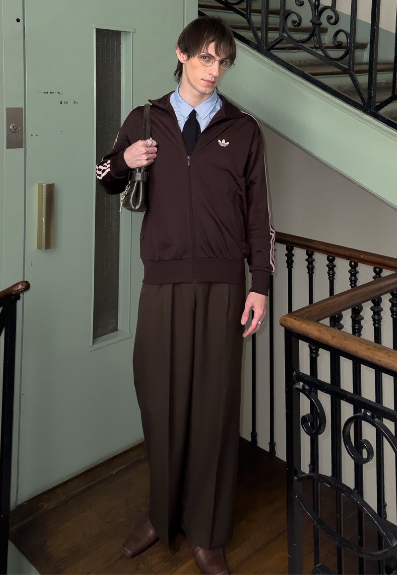 Jeune homme portant des lunettes, une veste Adidas marron, une chemise bleu clair, une cravate noire, un pantalon large foncé, tenant une petite pochette sur un palier d'escalier.