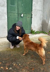 Osoba v černé péřové bundě a šedé čepici klečí vedle malého, červenohnědého psa s nadýchaným ocasem u zelených dveří.