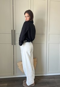 Zwarte linnen overhemd met knopen, witte wijde broek, beige instappers, natuurlijk geweven tas, staand tegen lichtgekleurde kasten.