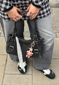 Black handbag with dual handles, adorned with a lace ribbon, a crocheted keychain, and metal hardware. Paired with oversized grey trousers.