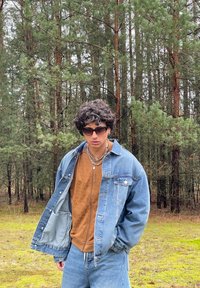 Giacca di jeans con lavaggio azzurro chiaro, indossata sopra un maglione marrone a fantasia. Jeans azzurri chiari e occhiali da sole completano il look. Sfondo di foresta.