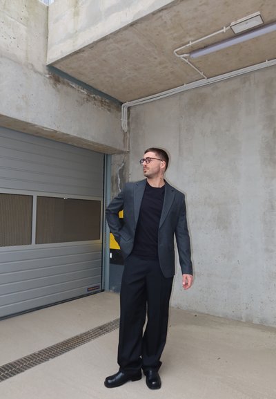 Hombre con traje oscuro y gafas de pie dentro de un garaje de hormigón cerca de una puerta metálica cerrada, mirando hacia su izquierda.