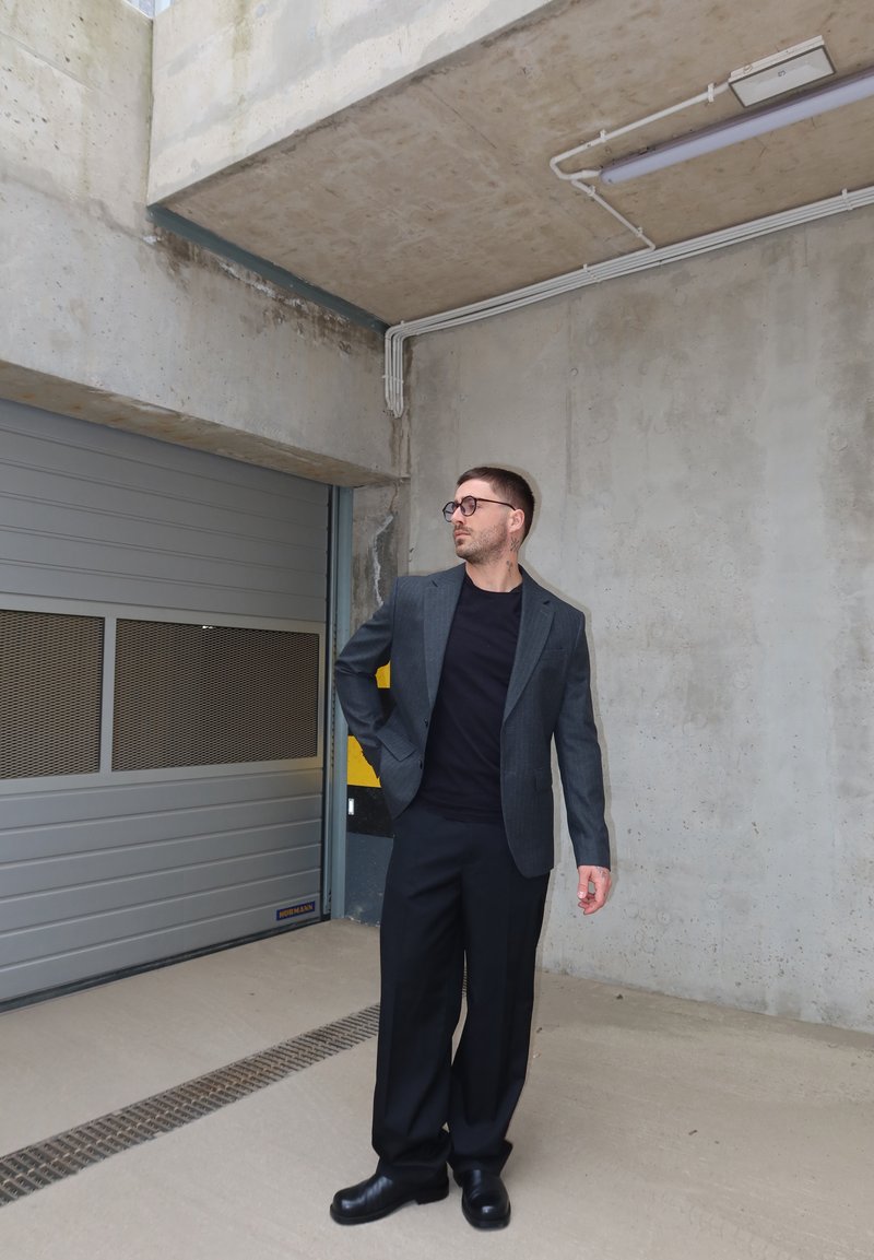 Man in donker pak en bril staat in een betonnen parkeergarage nabij een gesloten metalen deur en kijkt naar links.