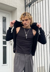 Jeune personne aux cheveux blonds bouclés en train d'enlever une veste noire, portant un haut noir sans manches à lacets sur les côtés, un pantalon plissé gris, et un collier argenté en forme de clé.