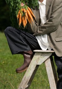 Personne assise sur une échelle en bois en plein air, portant des bottes marron, tenant une botte de carottes fraîches orange avec des fanes vertes.