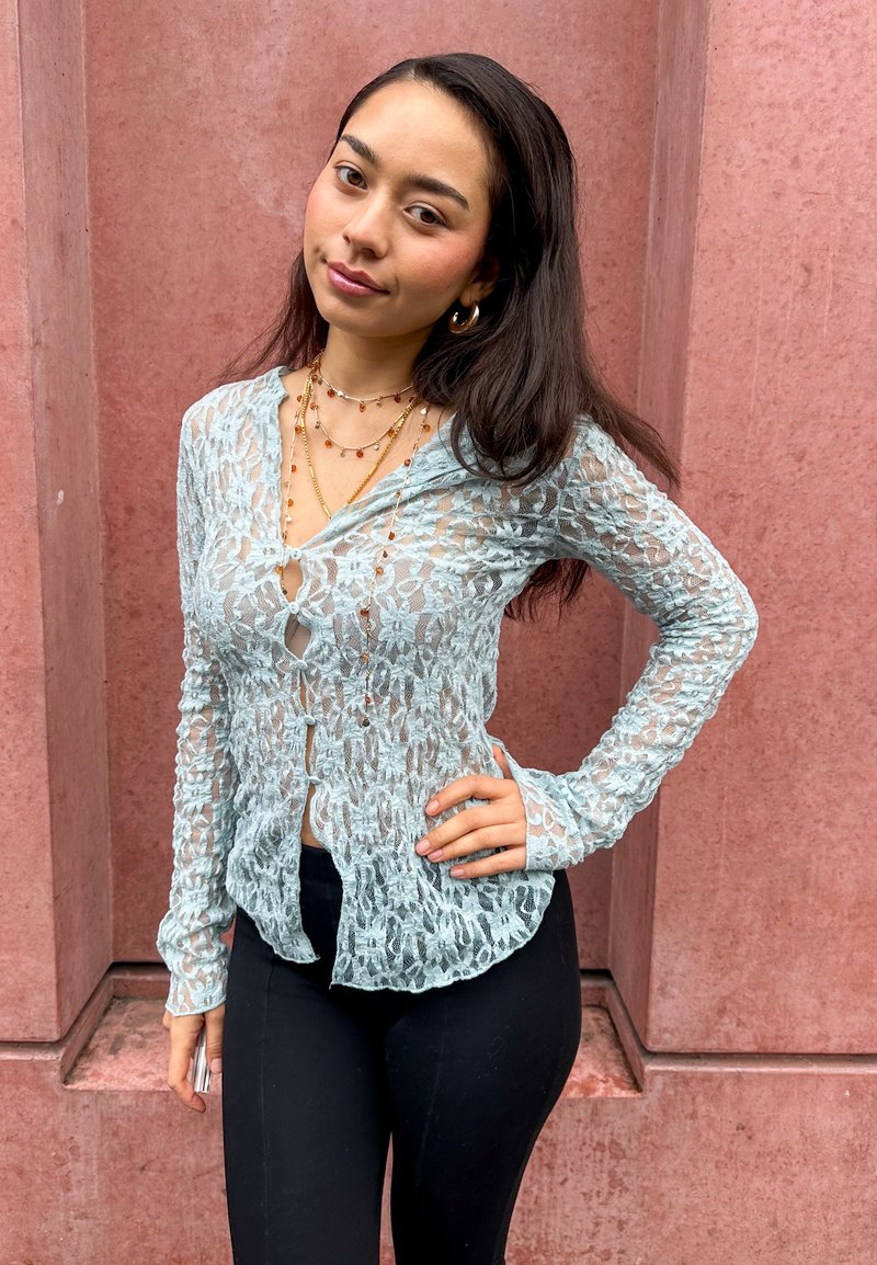 Cardigan en dentelle bleu clair à manches longues, motif floral texturé, avec une fermeture boutonnée à l'avant. Associé à un pantalon noir ajusté.