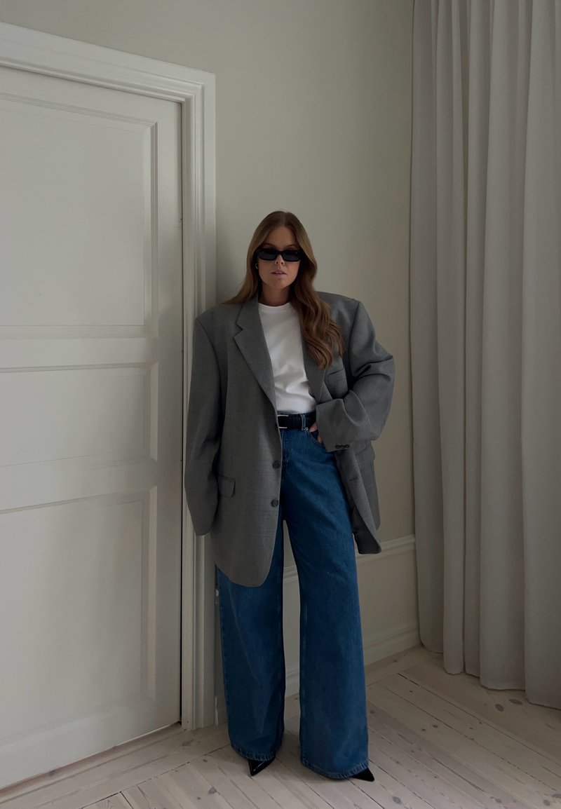 Femme aux longs cheveux bruns portant un blazer gris oversize, une chemise blanche, un jean large bleu, une ceinture noire, des lunettes de soleil, debout à l'intérieur près d'une porte fermée.