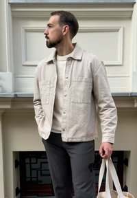 Lys beige knap-uppet jakke med to frontlommer, båret over en cremefarvet sweater, parret med grå bukser, holder solbriller og en tote taske.
